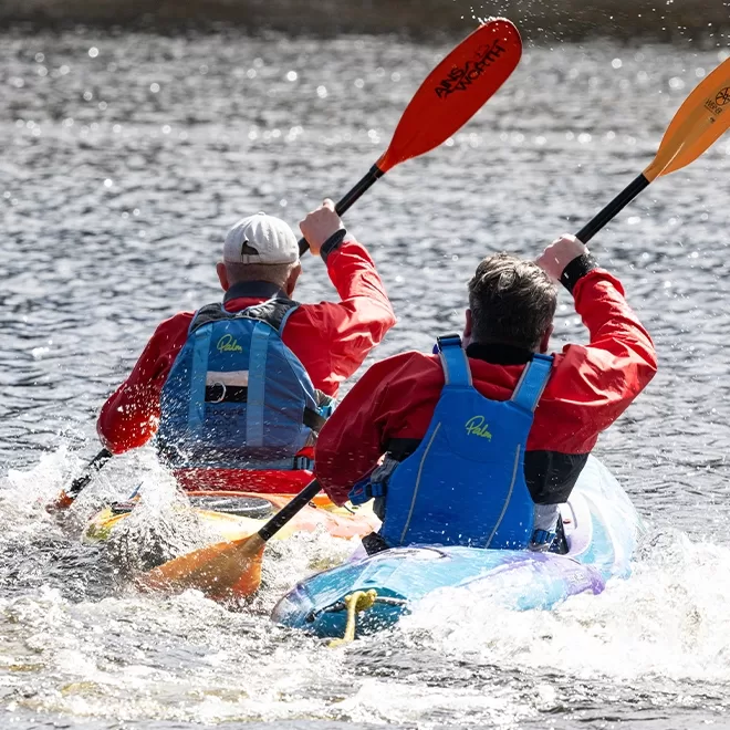 The back of two people wearing red rainproof jackets and life jackets, using paddles while kayaking in the loch - in Scotland.