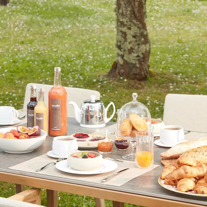 A table set for breakfast outdoors on a lawn. It features fruit, croissants, baguettes, butter, jam, juice, bottled drinks, a teapot, and cups. In the background are trees and grass scattered with white flowers.