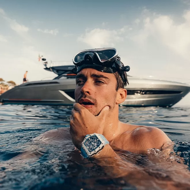Charles Leclerc in water wearing a diving mask on their head and a watch on their wrist, touching their chin. A sleek, modern boat is visible in the background against a partly cloudy sky.