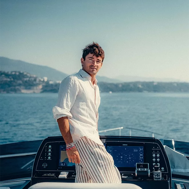 Charles Leclerc in a white shirt and striped pants stands at the helm of a boat. He gazes forward with a calm sea and distant hills in the background under a clear blue sky. The control panel of the boat is visible in front of him.