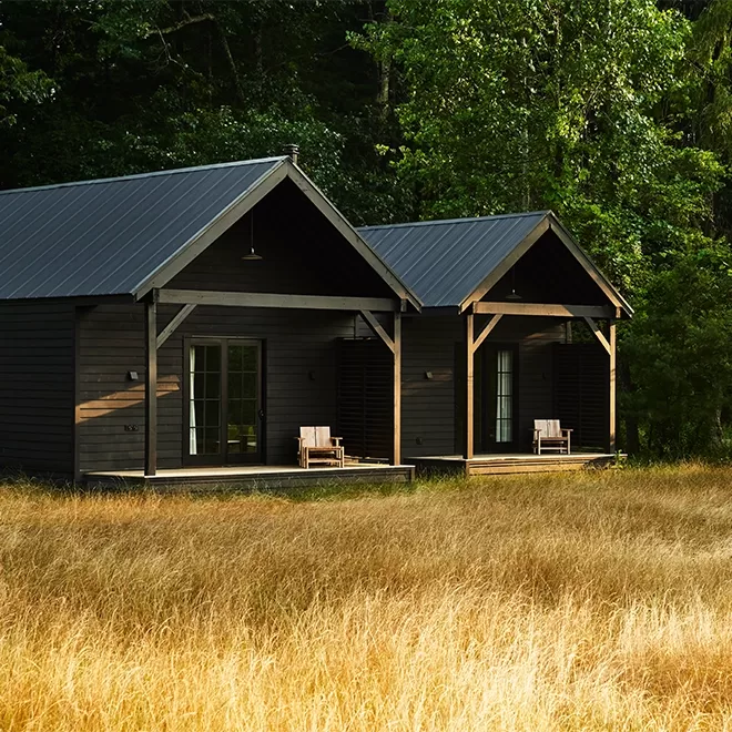 Two adjoining dark wooden cabins with steep roofs sit in a sunlit grassy field surrounded by dense green trees. Each cabin has a small porch with a wooden chair, and the ambiance is peaceful and rusticat INNESS,New York.