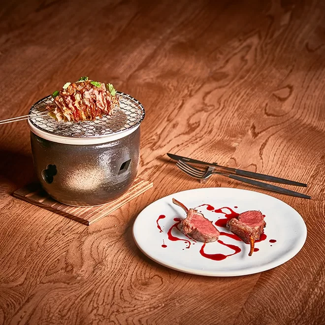 A small grill with sliced vegetables on a mesh surface is beside a white plate of two lamb chops garnished with sauce. The setup is on a wooden table, accompanied by a fork and knife at Humo, London.