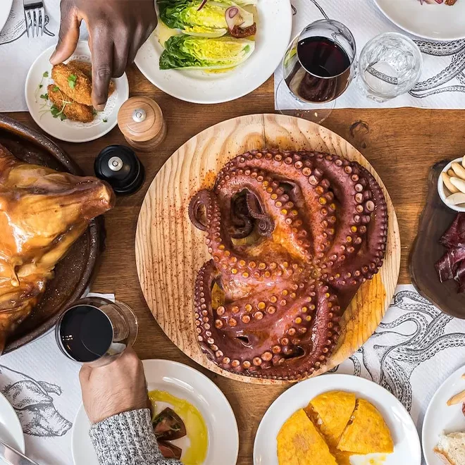 A wooden table is set with plates of food, including a large cooked octopus centerpiece, salad, bread, and various dishes. Several peoples hands are seen reaching for food. Glasses of red wine accompany the meal.