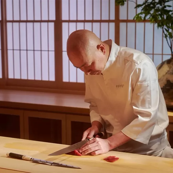 A focused chef in a white uniform is carefully slicing fish at a traditional wooden sushi counter, set against a serene backdrop of Japanese-style sliding doors at Sushi Kanesaka, London. His precise technique and dedication are evident in the quiet, intimate setting.