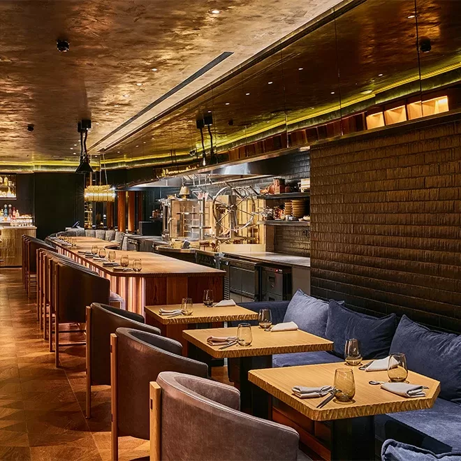 A modern, warmly lit restaurant interior featuring wooden tables set with glasses and cutlery. The seating includes upholstered chairs and a long bar counter in the background with decorative lighting and kitchen equipment visible at Humo, London.