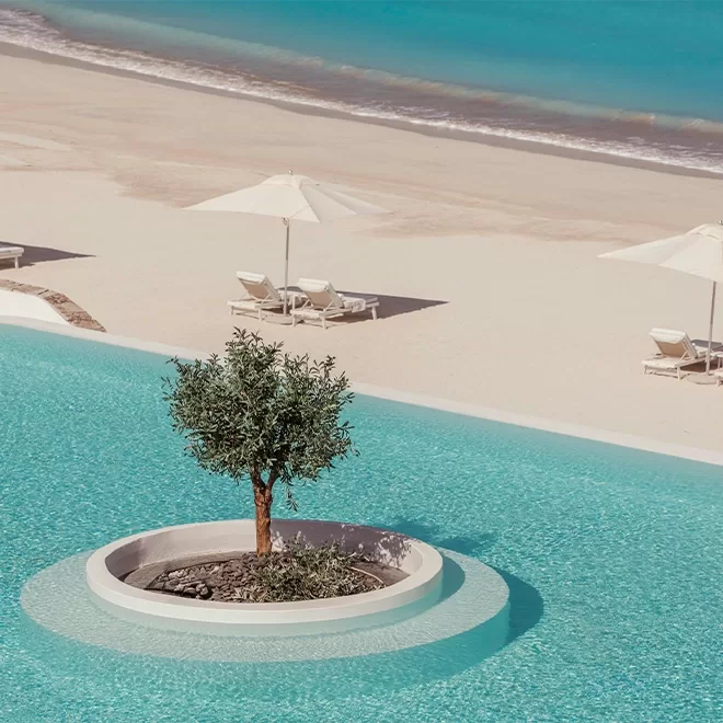 A serene beach scene features a sparkling turquoise pool in the foreground with a small tree planted on a circular island. In the background, sandy shores and clear blue sea are accompanied by white umbrellas and lounge chairs at Anantara Santorini Abu Dhabi Retreat, Abu Dhabi.