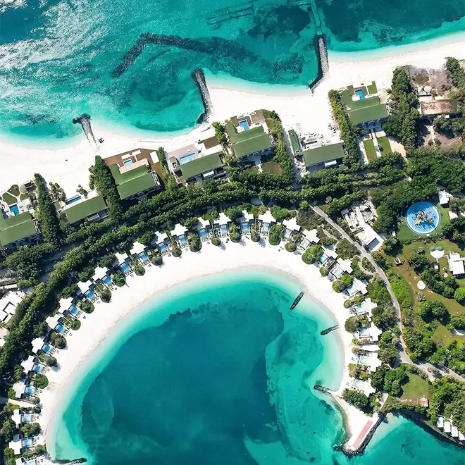 Aerial view of a tropical resort with white sandy beaches and clear turquoise water. Bungalows are arranged in a semi-circle along the shore, surrounded by lush greenery. Several piers extend into the ocean at  at Nurai Island Resort, Abu Dhabi.