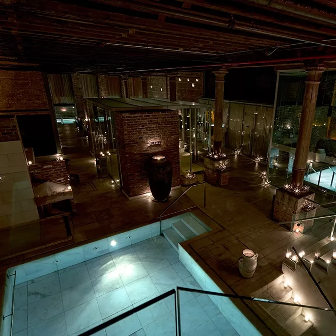 Dimly lit spa interior with multiple small pools and stone walls. Candles line the walkways, creating a serene atmosphere. Columns and soft lighting enhance the tranquil and rustic ambiance of the space.