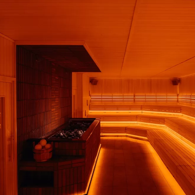 A warm, orange-lit sauna interior featuring tiered wooden benches along the walls, and a stone heating unit in the corner with a wooden bucket nearby. The rooms ambiance is cozy and inviting.