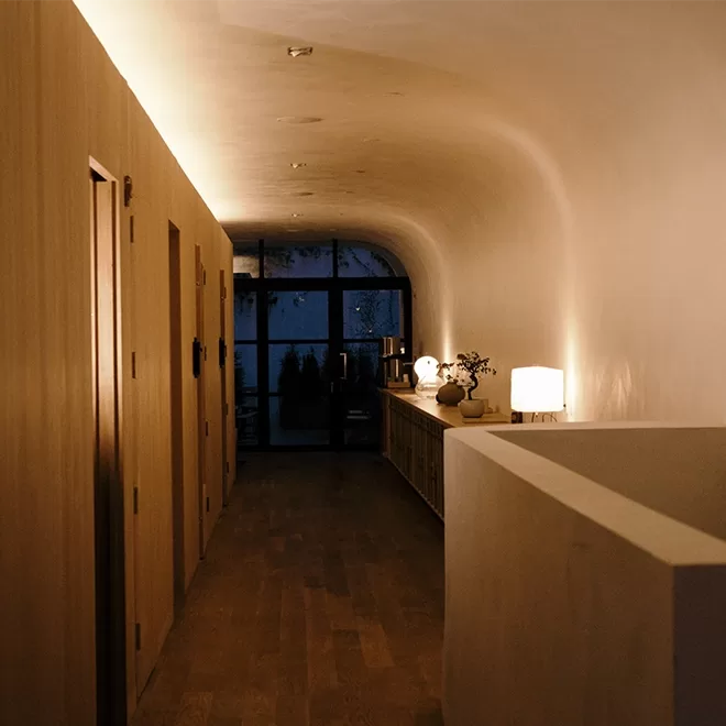 Dimly lit hallway with curved ceiling, featuring closed wooden doors on the left. A side table on the right holds small lamps casting a warm glow. At the end, a glass door reveals faint outdoor trees.