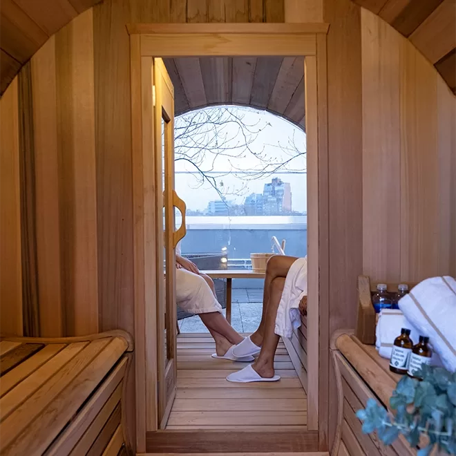 Two people in white robes relax in a wooden sauna. One persons legs, wearing white slippers, are visible through the saunas open door. Outside, buildings and a tree are visible under a cloudy sky. Sauna supplies sit on a shelf inside.