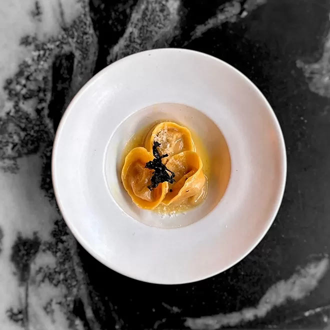 A white plate holds three neatly arranged pieces of stuffed pasta in a light sauce, topped with a garnish, on a marbled black and white background at Angelina, Dalston.