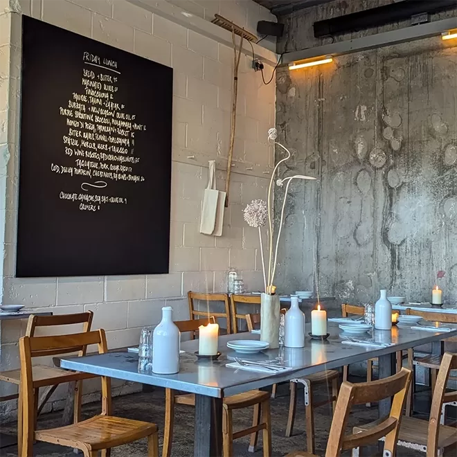 A cosy restaurant interior with wooden chairs and tables set with white plates, jars, and candles. A large blackboard on the wall displays a menu. The decor includes rustic gray walls and minimalistic art pieces at Jolene, Newington Green.