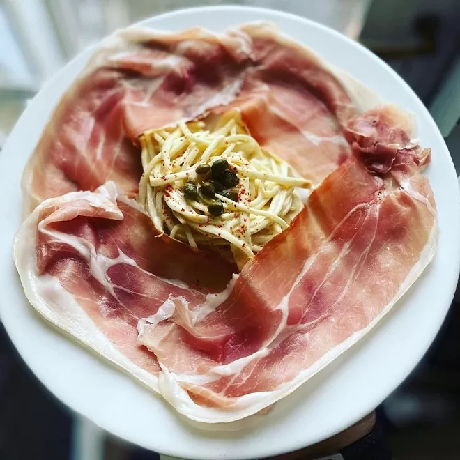 Plate of thinly sliced prosciutto arranged around a central portion of pasta topped with green capers. The dish is presented on a white plate at Bouchon Racine, Farringdon.