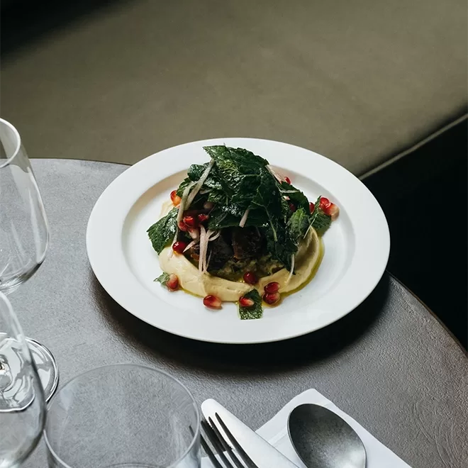 A plate of gourmet food featuring a dish with leafy greens, pomegranate seeds, and a dark central piece, presented on a white plate atop a gray table. Nearby are empty wine glasses, a fork, spoon, and a white napkin at OMA, Borough Market.