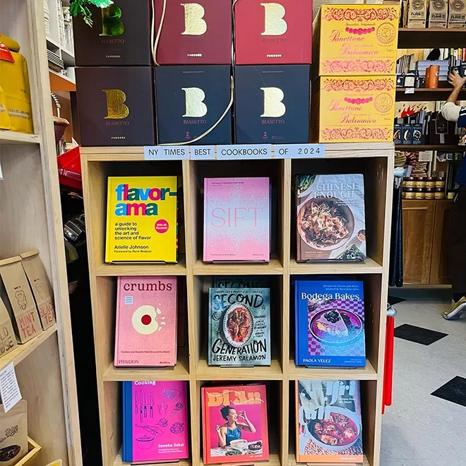 A bookshelf at Archestratus, Greenpoint in NYC displays, various cookbooks, including Flavorama, Sift, Chinese Takeout Made Easy, Crumbs, The Second Generation, Bodega Bakes, and Diaspora Diner. Boxes labeled Ovenbird and Panettone are on top.