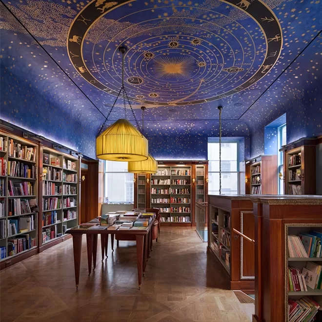 A library room inside Albertine, Upper East Side in NYC with, bookshelves lining the walls and a wooden floor. The ceiling is painted with a celestial design featuring constellations. Three large, yellow pendant lights hang over a central long table filled with books.