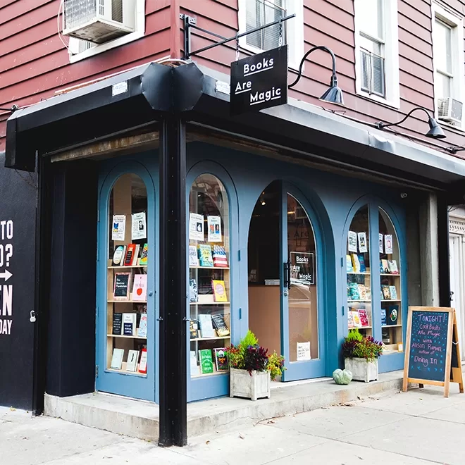 A corner bookstore with a sign reading Books Are Magic in NYC. The shop has large arched windows displaying a variety of books. A chalkboard easel stands outside with handwritten notes. The building exterior is red with blue accents.