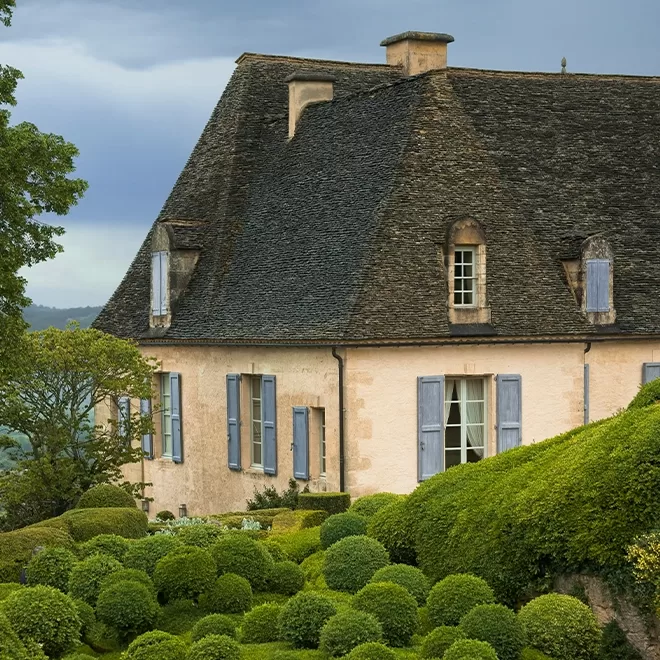 The charming house - Château de Marqueyssac, with a steep, dark slate roof, cream-colored walls, and blue shutters. It is surrounded by neatly trimmed bushes and lush greenery under a cloudy sky.
