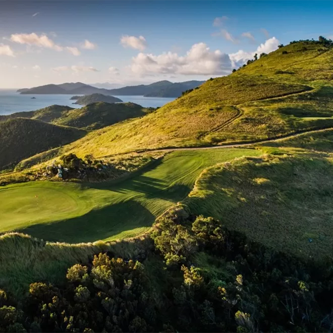 Aerial view of a golf course on a lush, green hillside with winding paths. Surrounding hills and distant islands are visible under a partly cloudy sky. The landscape overlooks a calm, blue ocean.