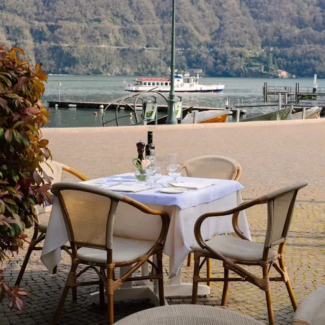 An outdoor dining table set with white linens and glassware is surrounded by four chairs near a waterfront, with a boat and hilly landscape visible across the water in the background.