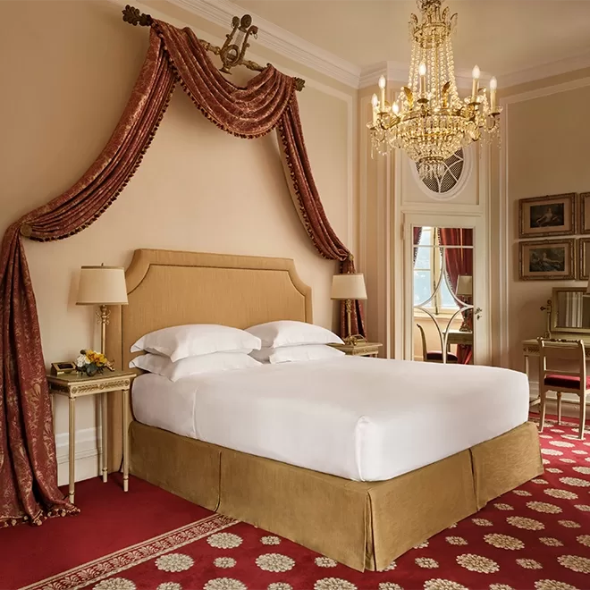 Elegant hotel room with a large bed, gold headboard, draped red curtains, ornate chandelier, classic lamps, a mirror, and antique-style furniture on a red patterned carpet. The décor is luxurious and traditional.