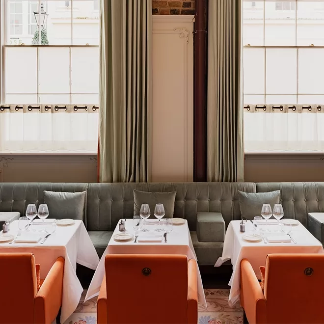 A stylish restaurant dining area with sage green velvet banquette seating, orange upholstered chairs, white tablecloths, set tables with glassware, and large windows with light green curtains.