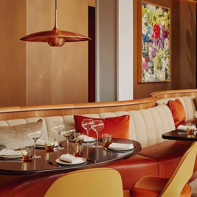 A modern restaurant interior with a cushioned beige banquette, set dining tables, amber and red accents, a copper hanging lamp, and a colorful floral painting on the wall.