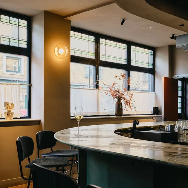 A modern bar interior with a curved marble countertop, black chairs, a glass of white wine, and a vase with twigs. Large windows let in natural light, and the walls are decorated in neutral tones.
