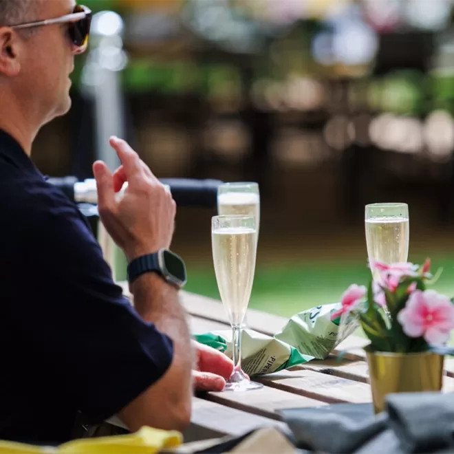 A person wearing sunglasses and a smartwatch sits at an outdoor wooden table with two glasses of champagne, a small bouquet of pink flowers, and snack bags. The background is blurred with greenery.