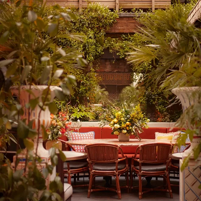 A cozy outdoor dining area with a round table, red cushioned chairs, and floral centerpieces, surrounded by lush green plants and vines on brick walls.