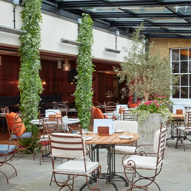 A stylish outdoor restaurant patio with round wooden tables, cushioned metal chairs, tall green plants, and a glass roof. Some tables have menus, and large potted plants add greenery to the elegant space.