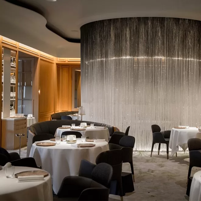 Elegant restaurant interior with round tables covered in white tablecloths, dark chairs, and a dramatic curved wall featuring shimmering strings or beads, softly lit to create a luxurious atmosphere.