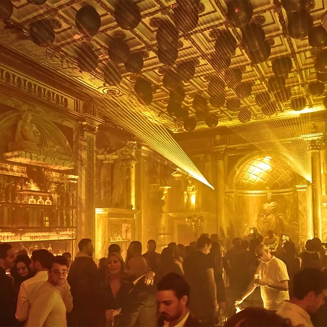 A crowded, opulent nightclub with golden lighting, ornate statues, and a decorated ceiling. People are socializing, some holding drinks, under dramatic beams of light. The atmosphere is lively and elegant.