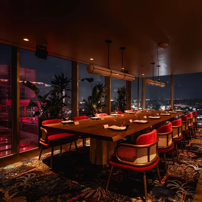 A dimly lit upscale dining room with a long wooden table set for a meal, surrounded by red and cane chairs, large windows showing a city view at night, and plants adding a cozy ambiance.