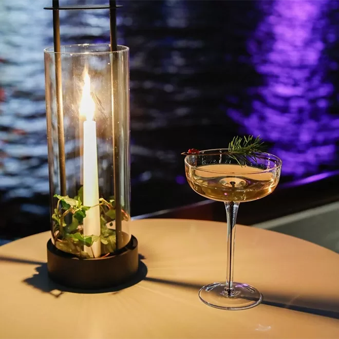 A lit candle in a glass holder sits on a table next to a cocktail in a coupe glass, garnished with herbs. In the background, water reflects purple and blue lights.