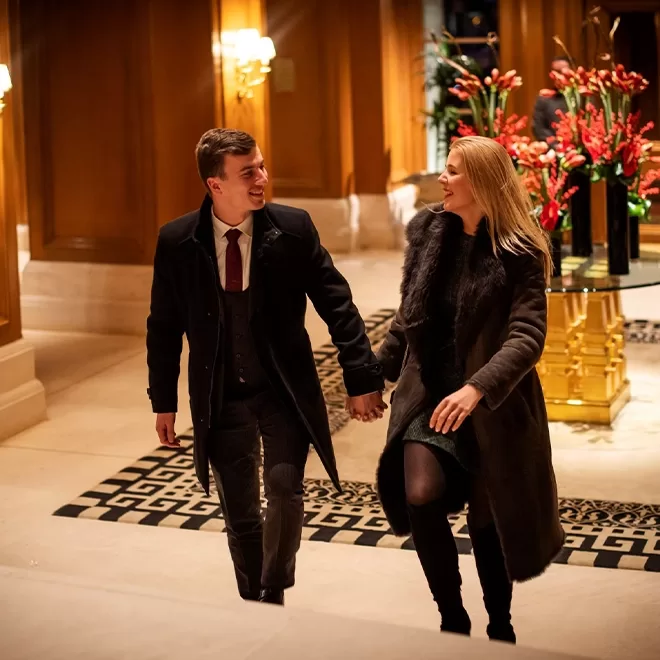 A smiling man and woman, both dressed in formal winter clothes, hold hands while walking up elegant, marble stairs in a luxurious, warmly lit interior decorated with large floral arrangements.