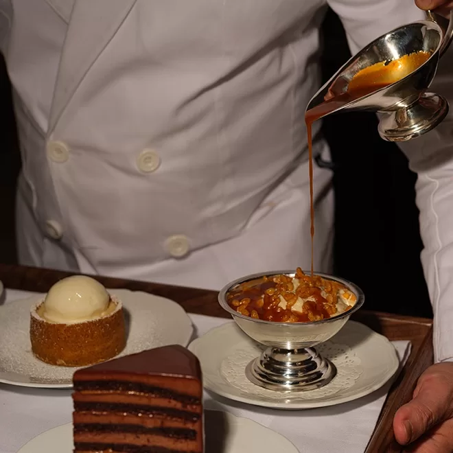 A person in a white chefs coat pours caramel sauce over a sundae in a silver dish, with chocolate cake and a dessert topped with a scoop of ice cream on a tray nearby.