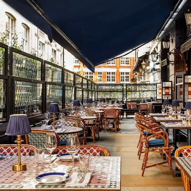 Outdoor restaurant seating with patterned tables, rattan chairs, blue-shaded lamps, wine glasses, and tableware. The area is covered by a large awning with city buildings visible in the background.