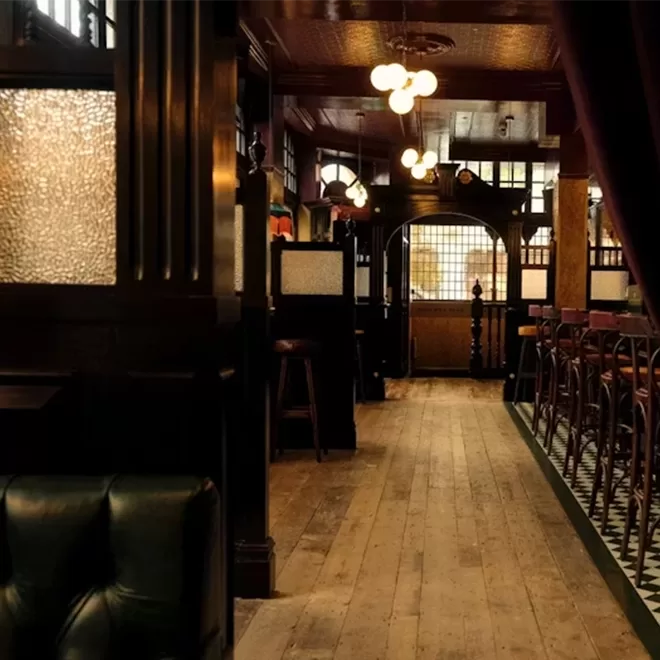 A warmly lit, vintage-style pub interior with wooden floors, leather seating, high bar stools, checkered tile, and frosted glass partitions, creating a cozy, inviting atmosphere.