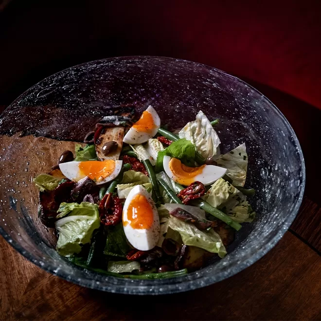 A glass bowl filled with a salad of lettuce, green beans, sun-dried tomatoes, olives, and quartered soft-boiled eggs with runny yolks, set on a wooden table with a dark red background.