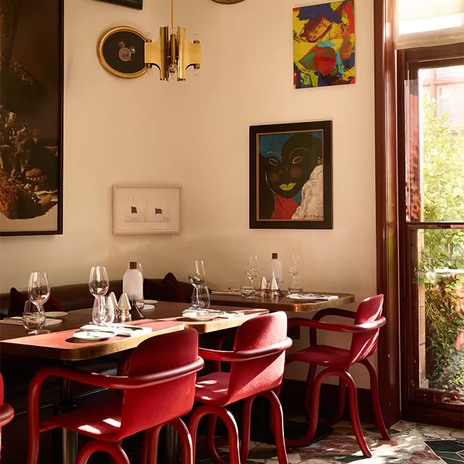 A cozy restaurant dining area with red chairs, set tables with glassware, and eclectic artwork on cream-colored walls. Sunlight streams in through a tall window beside the tables.