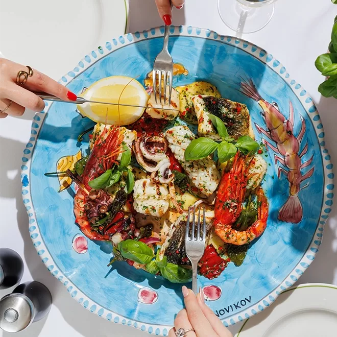 A colorful seafood dish with shrimp, fish, and vegetables is served on a blue decorative plate. Two hands are reaching in with forks, and one hand is holding a lemon wedge above the plate. Fresh basil garnishes the dish.
