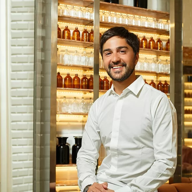 A man with short dark hair and a beard, wearing a white button-up shirt, smiles while sitting in front of shelves filled with amber and black bottles in a brightly lit room.
