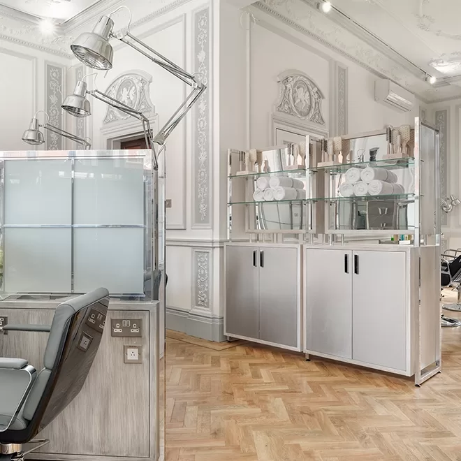 A modern, elegant salon with ornate white walls, wooden herringbone floor, glass display shelves holding towels and products, and a stylish salon chair near a frosted glass divider.