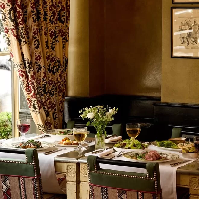 A cozy restaurant table set for four with white linens, elegant place settings, plates of food, glasses of wine and water, and a small vase of flowers, surrounded by patterned curtains and framed art on the wall.