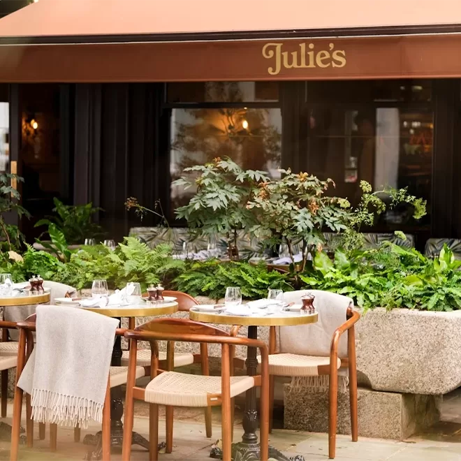 Outdoor dining area at Julie’s restaurant with wooden tables, chairs draped with light blankets, set with glassware and napkins, surrounded by lush green plants, under an orange awning displaying the restaurant’s name.