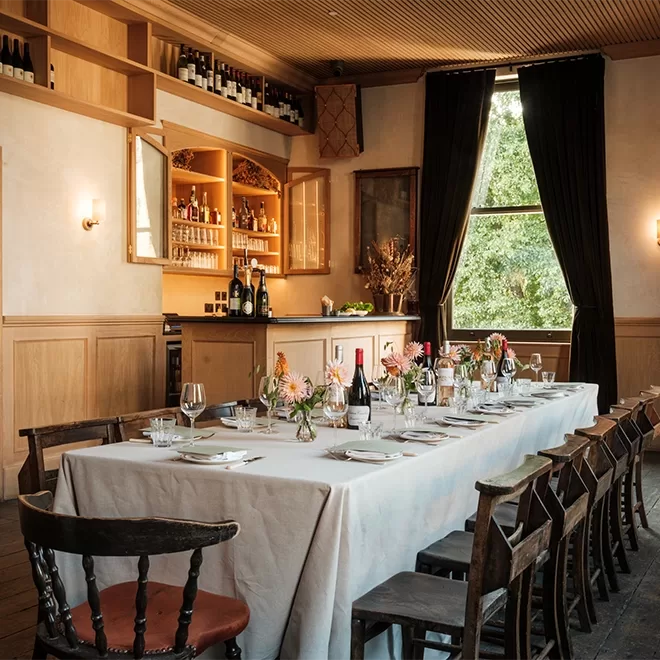 A long dining table with a white tablecloth is set with plates, glasses, and small floral arrangements in a warmly lit room with wooden chairs, large windows, and shelves stocked with bottles.