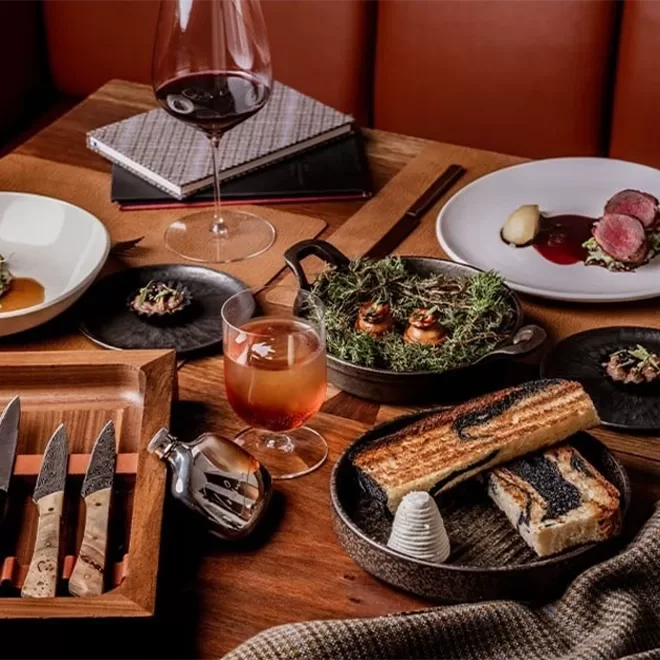 A restaurant table set with gourmet dishes, toasted bread with butter, assorted meats, creative appetizers, a glass of red wine, a cocktail, and elegant cutlery, creating a sophisticated fine dining atmosphere.