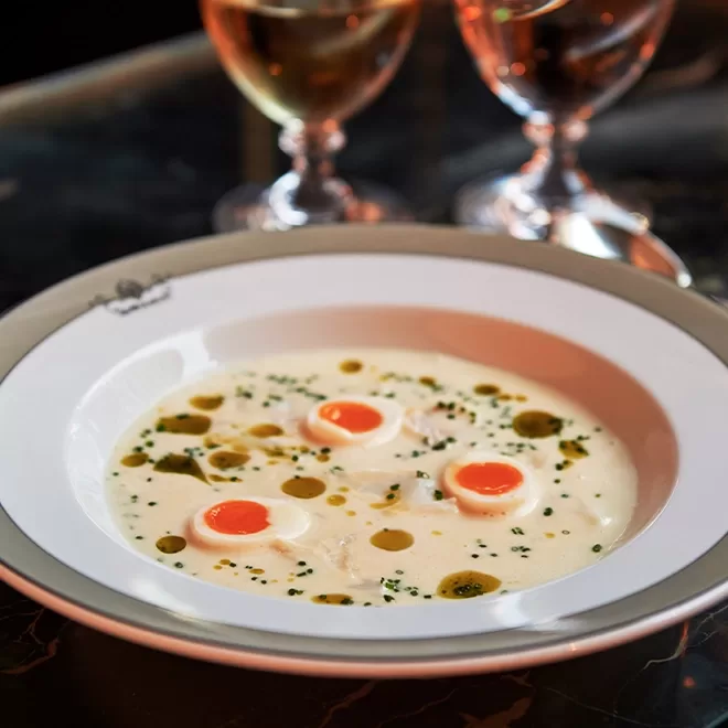 A bowl of creamy white soup garnished with three soft-boiled eggs, herbs, and drops of oil, served on a patterned plate. Two glasses of wine are blurred in the background.